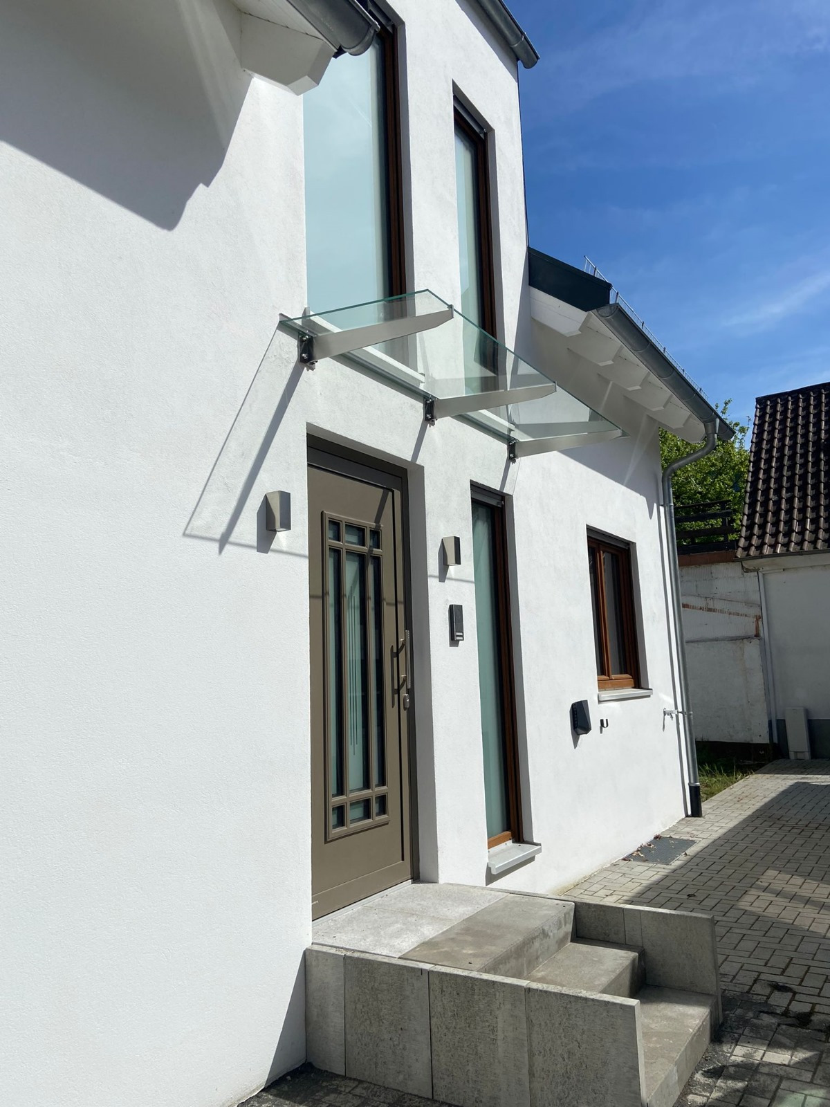 Wejście do nowoczesnego domu z szarobrązowymi drzwiami i szklanym zadaszeniem wspartym na metalowych wspornikach, oświetlone bocznym światłem słonecznym, z widocznymi schodami wejściowymi...