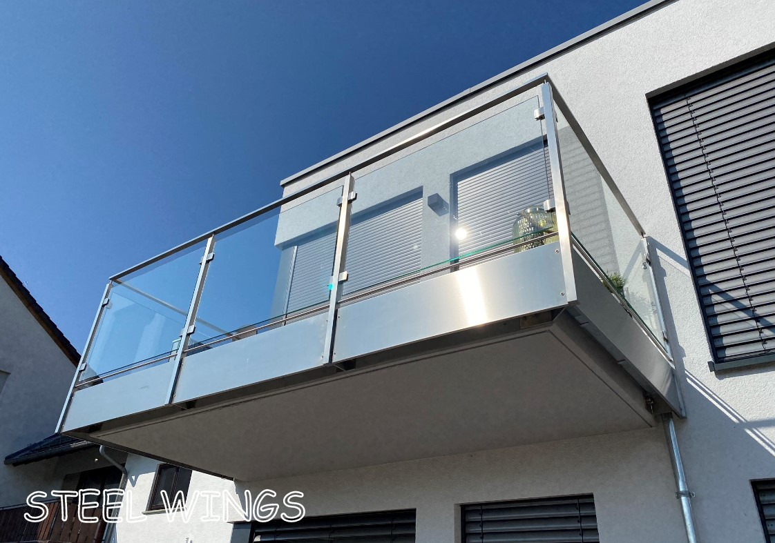 Nowoczesny balkon z przeszkloną, nierdzewną barierką na tle błękitnego nieba i fasady budynku z roletami.