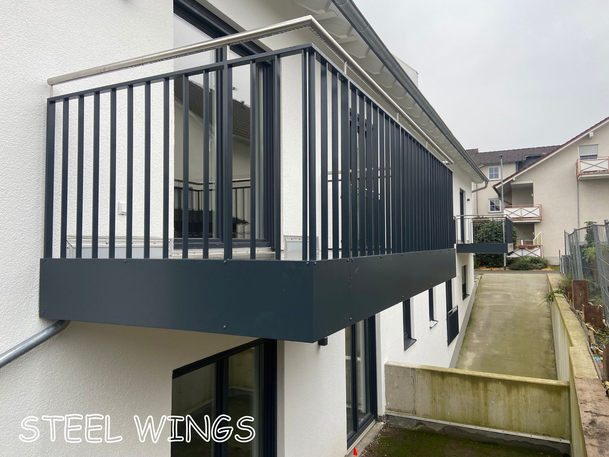 Nowoczesny balkon z czarną, metalową balustradą o pionowych szczeblach i nierdzewną poręczą, zamontowany przy białym budynku z dużymi oknami.