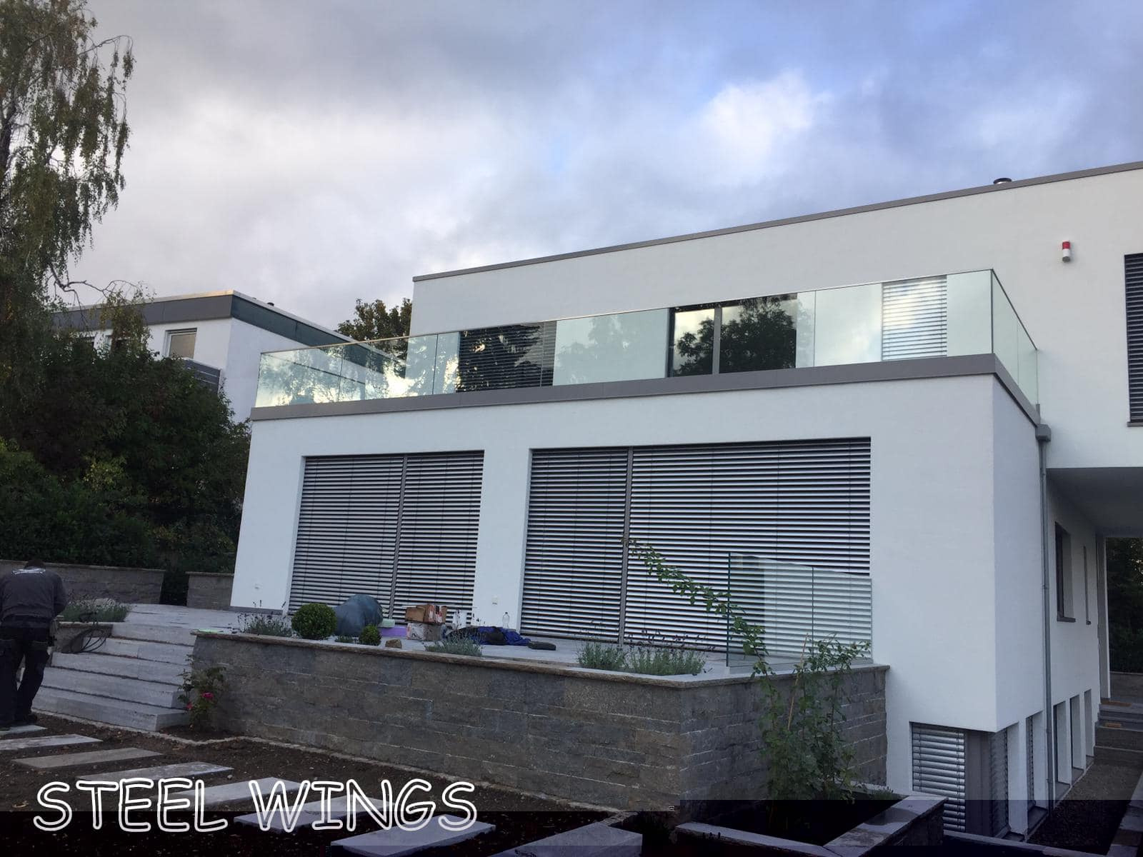 Nowoczesny dom z balkonem zabezpieczonym szklaną konstrukcją, widoczne żaluzje w oknach i fragment ogrodu z kamiennym murkiem.