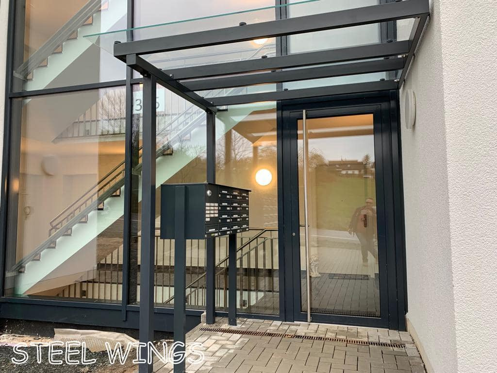 Nowoczesne wejście do budynku z przeszklonymi drzwiami i zadaszeniem, skrzynki pocztowe na metalowym stelażu, widok na schody wewnętrzne przez duże okno.