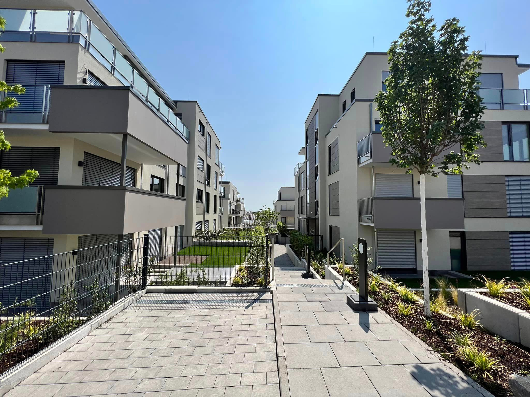 Nowoczesne osiedle z balkonami z przeszklonymi barierkami, niskimi metalowymi ogrodzeniami i brukowanym chodnikiem w słoneczny dzień.