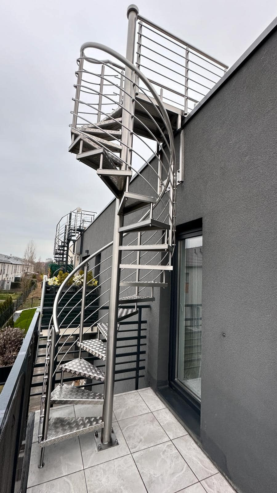 Zewnętrzne schody spiralne ze stali nierdzewnej z balustradą, prowadzące na dach budynku z szarą elewacją i widocznym oknem.