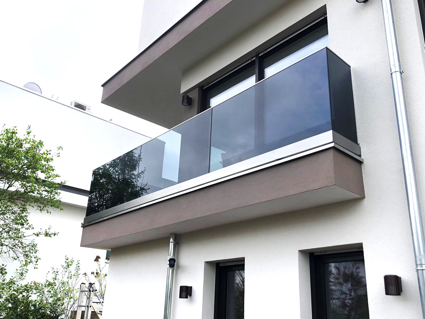 Nowoczesny balkon z szklaną balustradą na tle minimalistycznej elewacji budynku, widoczne odbicia drzew w szkle.