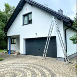 Dwukondygnacyjny dom jednorodzinny z garażem w trakcie prac wykończeniowych elewacji, z rozstawioną aluminiową drabiną opartą o ścianę, widoczne narzędzia i wiadro na podjeździe z kostki brukowej.