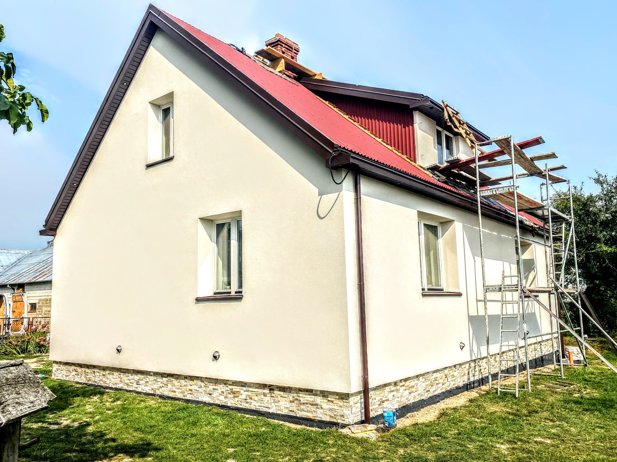 Dom jednorodzinny w trakcie prac elewacyjnych: nowa czerwona blacha na dachu, świeżo otynkowana i pomalowana ściana w kolorze kremowym, częściowo wykończony cokół z kamienia dekoracyjnego...