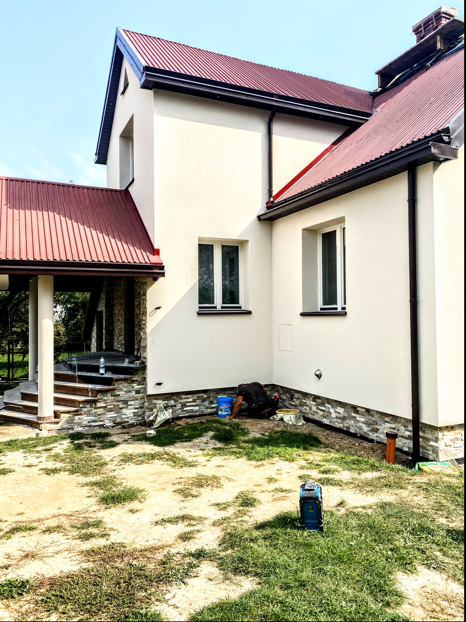 Pracownik w trakcie prac wykończeniowych przy elewacji domu jednorodzinnego, widoczny fragment ocieplenia i nowej elewacji, narzędzia pracy oraz agregat malarski.