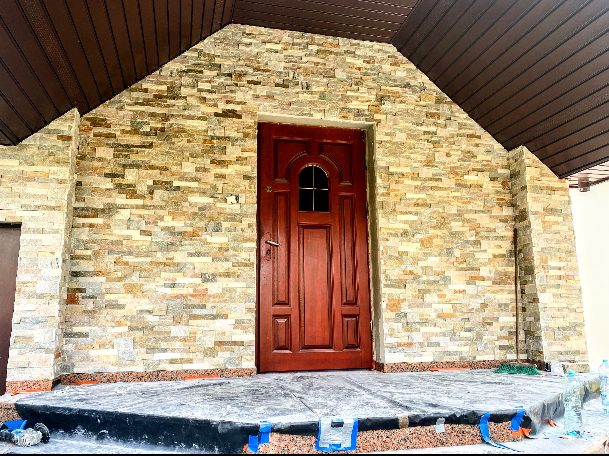 Wejście do domu z nową elewacją z kamienia dekoracyjnego i ciemnoczerwonymi drzwiami, zabezpieczone folią budowlaną, widoczna szczotka i butelki z wodą.