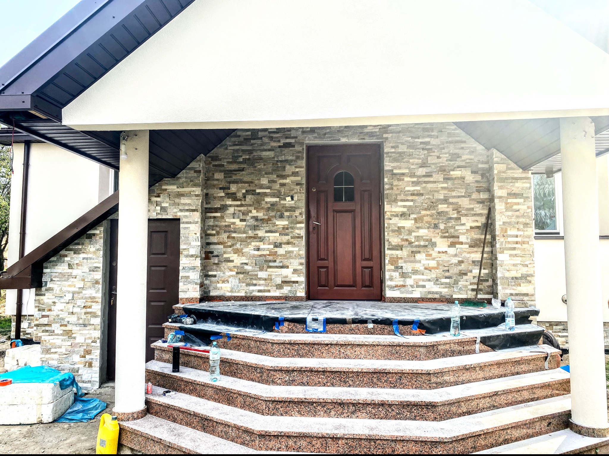 Wejście do domu z brązowymi drzwiami i schodami z czerwonego granitu, elewacja wykończona kamieniem dekoracyjnym, widoczne materiały budowlane i butelki z wodą na schodach.