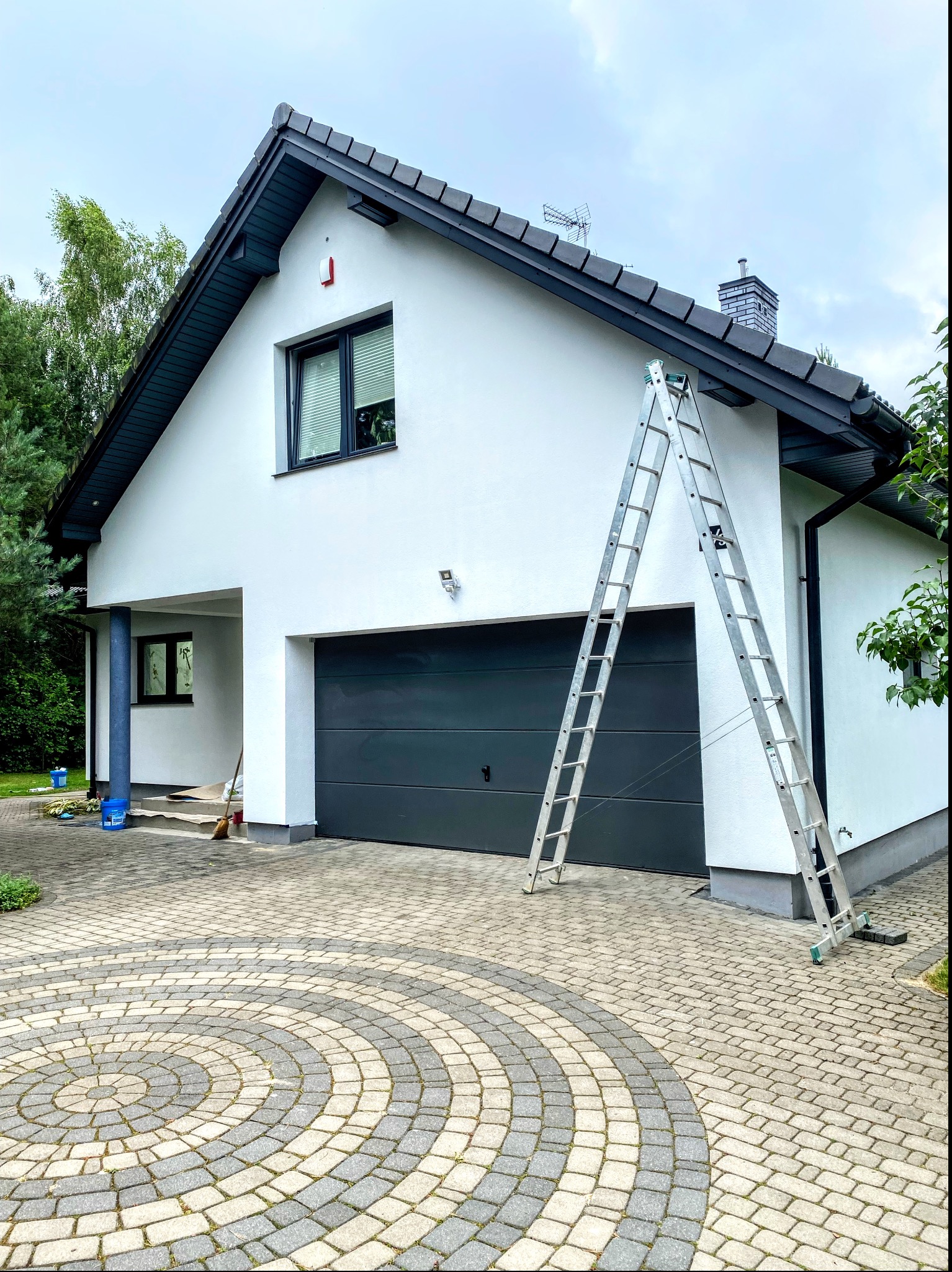 Dwukondygnacyjny dom jednorodzinny z garażem w trakcie prac wykończeniowych elewacji, z rozstawioną aluminiową drabiną opartą o ścianę, widoczne narzędzia i wiadro na podjeździe z kostki brukowej.