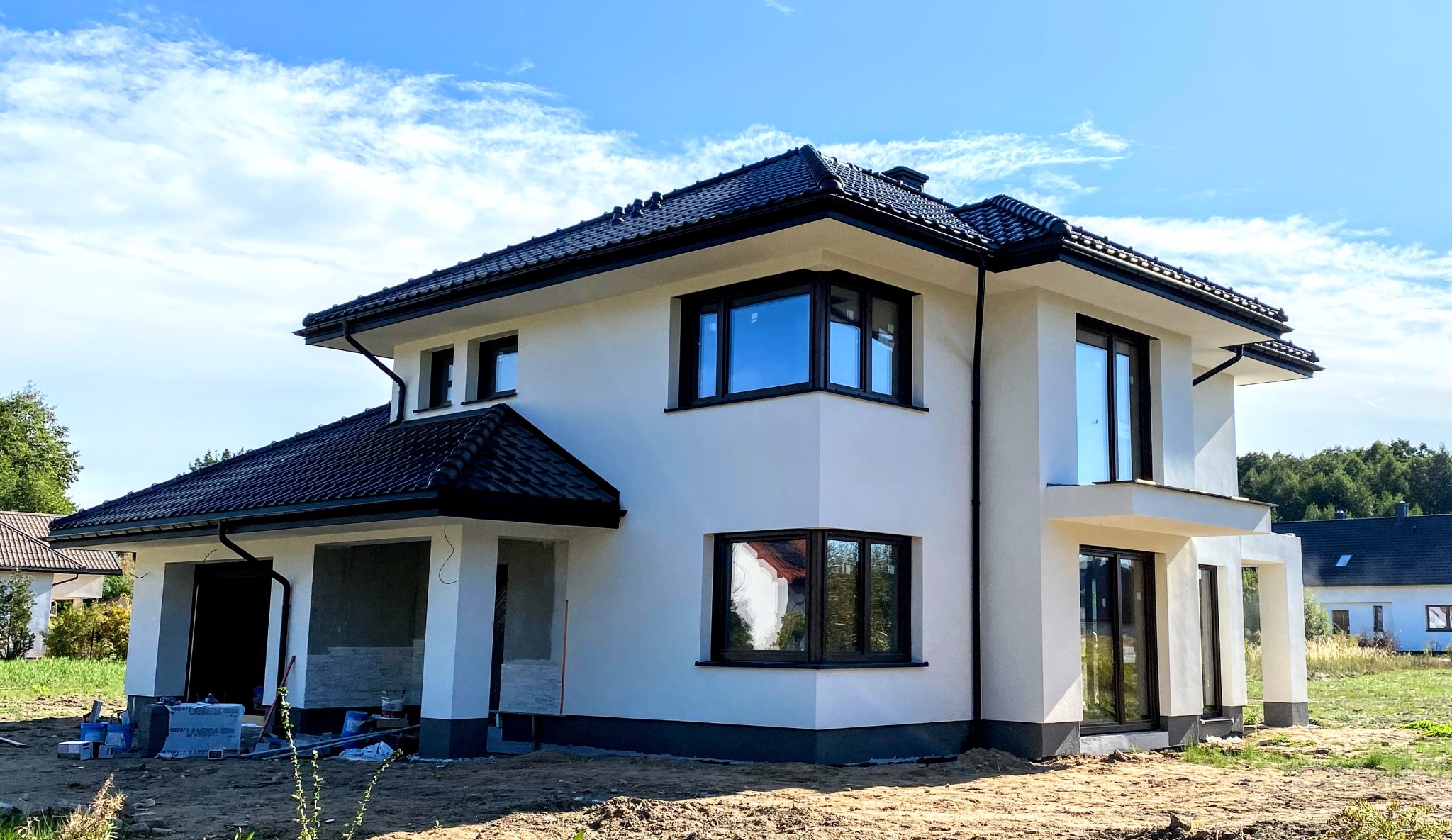 Dwukondygnacyjny dom jednorodzinny z białymi ścianami i czarną stolarką okienną, w trakcie prac wykończeniowych elewacji, widoczne materiały budowlane przy garażu.