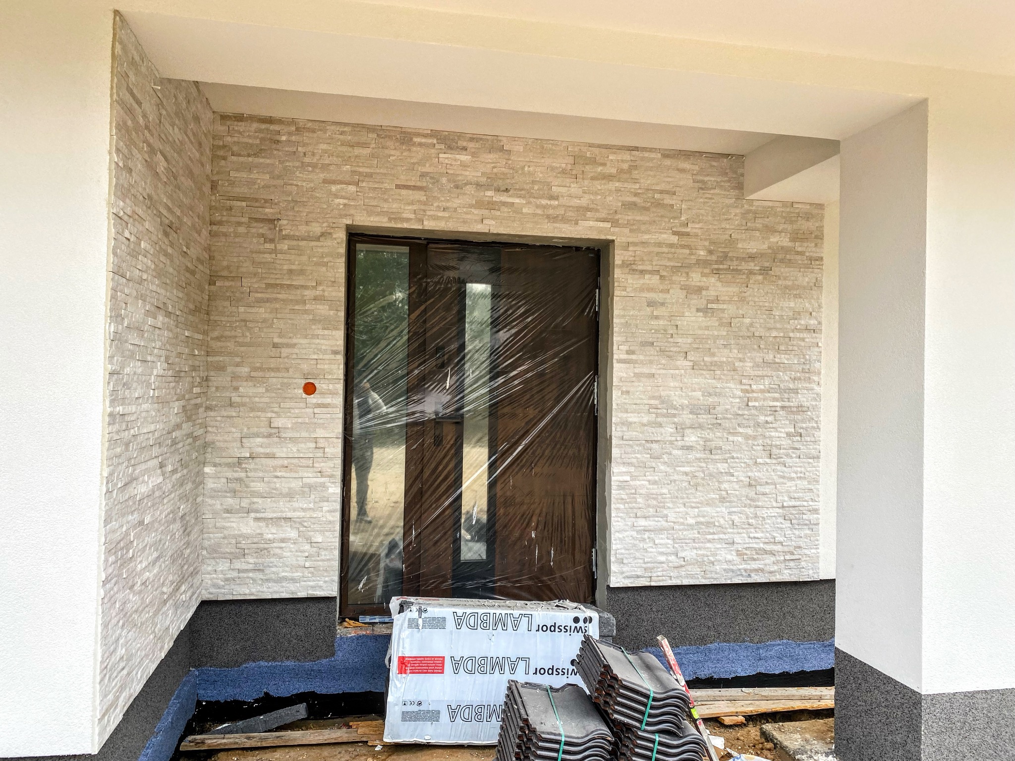 Wejście do domu z elewacją wykończoną dekoracyjnym kamieniem, zabezpieczone folią drzwi, ułożone dachówki i styropian czekające na montaż.