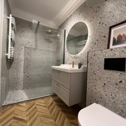 Rinaluck s.z o.o. - Nowoczesna łazienka w Gdyni z kabiną prysznicową, umywalką z szafką w kolorze gołębim, okrągłym lustrem z podświetleniem LED i płytkami imitującymi lastryko na ścianach oraz drewnopodobną podłogą...