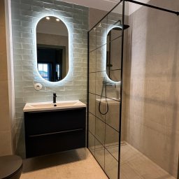 Rinaluck s.z o.o. - Nowoczesna łazienka z umywalką na szafce w kolorze czarnym, podświetlanym lustrem owalnym i kabiną prysznicową z czarnymi ramami na jasnych płytkach.