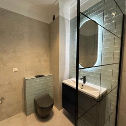 Rinaluck s.z o.o. - Nowoczesna łazienka z szarobeżowymi płytkami, podwieszaną toaletą i umywalką z czarną szafką, akcentem są niebieskie płytki za toaletą i podświetlane lustro w czarnej ramie.