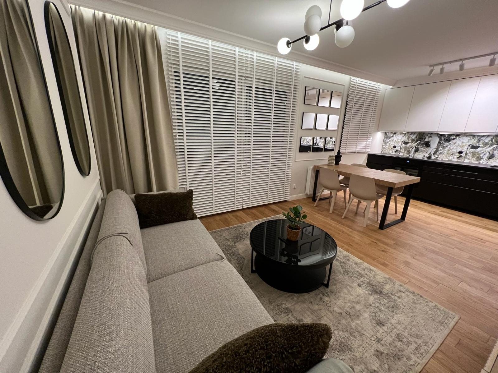 Jasne, nowocześnie urządzone wnętrze salonu z sofą w odcieniach szarości, czarnym stolikiem kawowym z rośliną, drewnianą podłogą i zasłonami w kolorze beżowym, widok na jadalnię z drewnianym stołem...