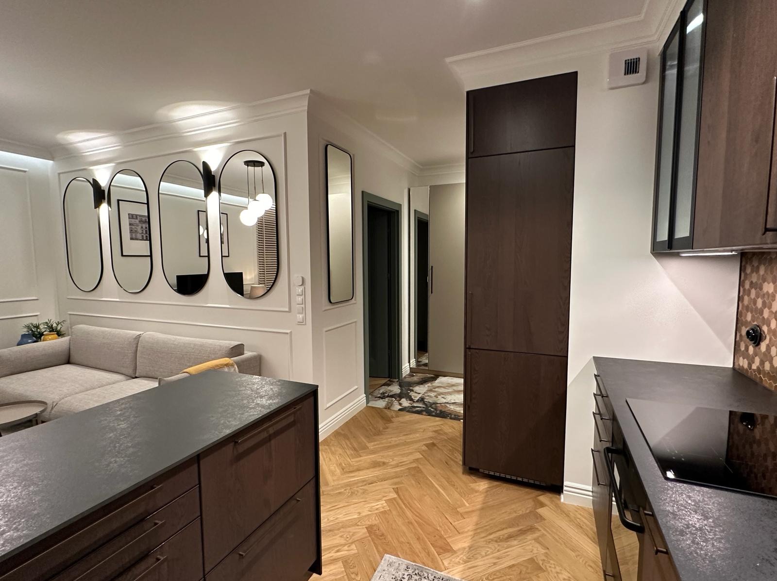 Nowoczesne wnętrze: parkiet w jodełkę, ściana z owalnymi lustrami, szara sofa, ciemne meble kuchenne z blatem imitującym kamień i mozaiką na ścianie.