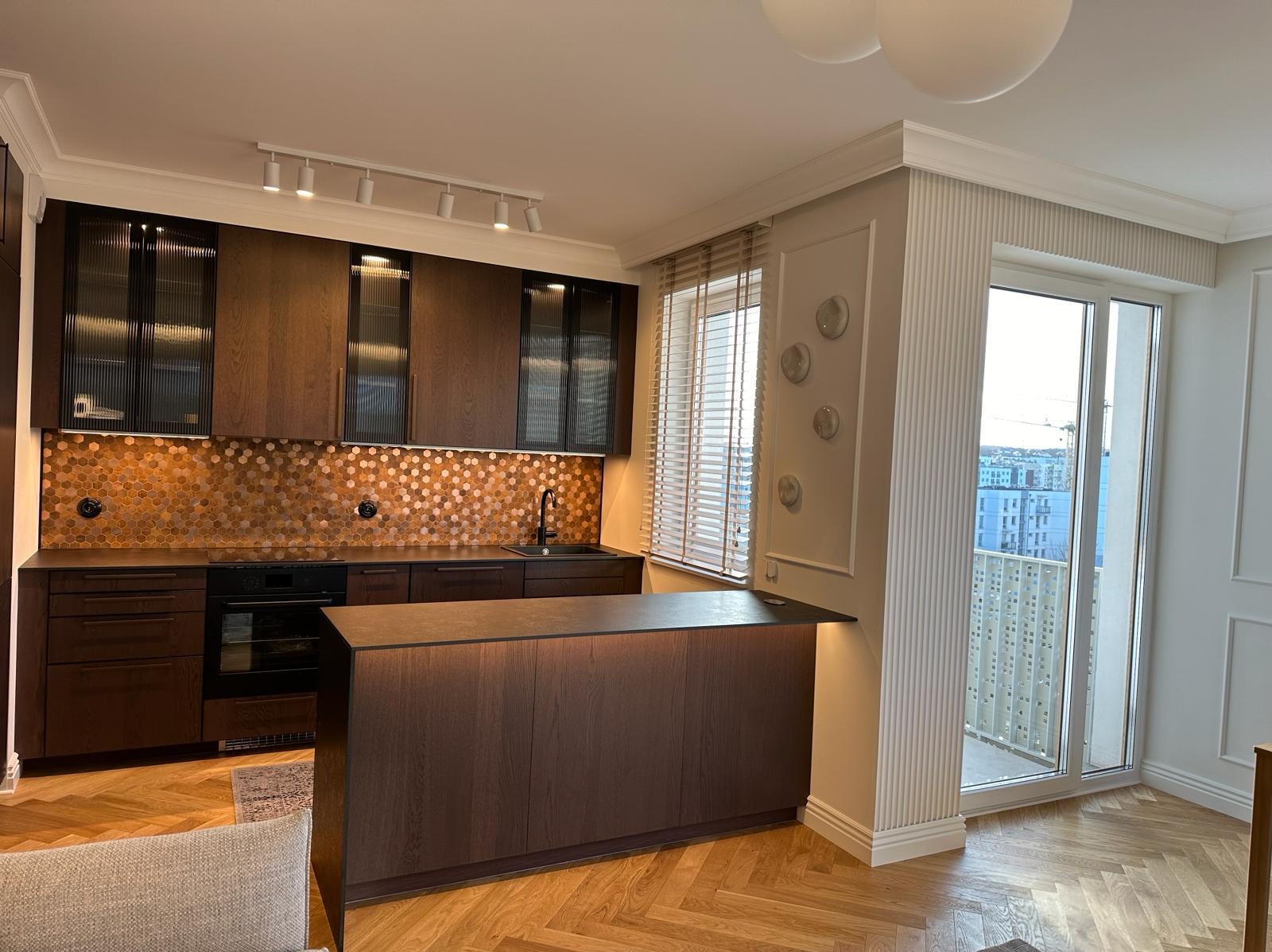 Nowoczesna kuchnia z ciemnymi drewnianymi szafkami, heksagonalnymi płytkami na ścianie i wyspą z podświetleniem LED, parkiet w jodełkę na podłodze, widok na miasto z balkonu.