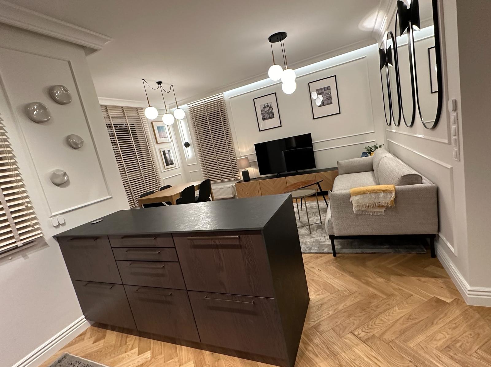 Nowoczesne wnętrze mieszkania z drewnianą podłogą w jodełkę, wyspą kuchenną z ciemnego drewna i szarym narożnikiem z żółtym pledem, oświetlone kulistymi lampami.