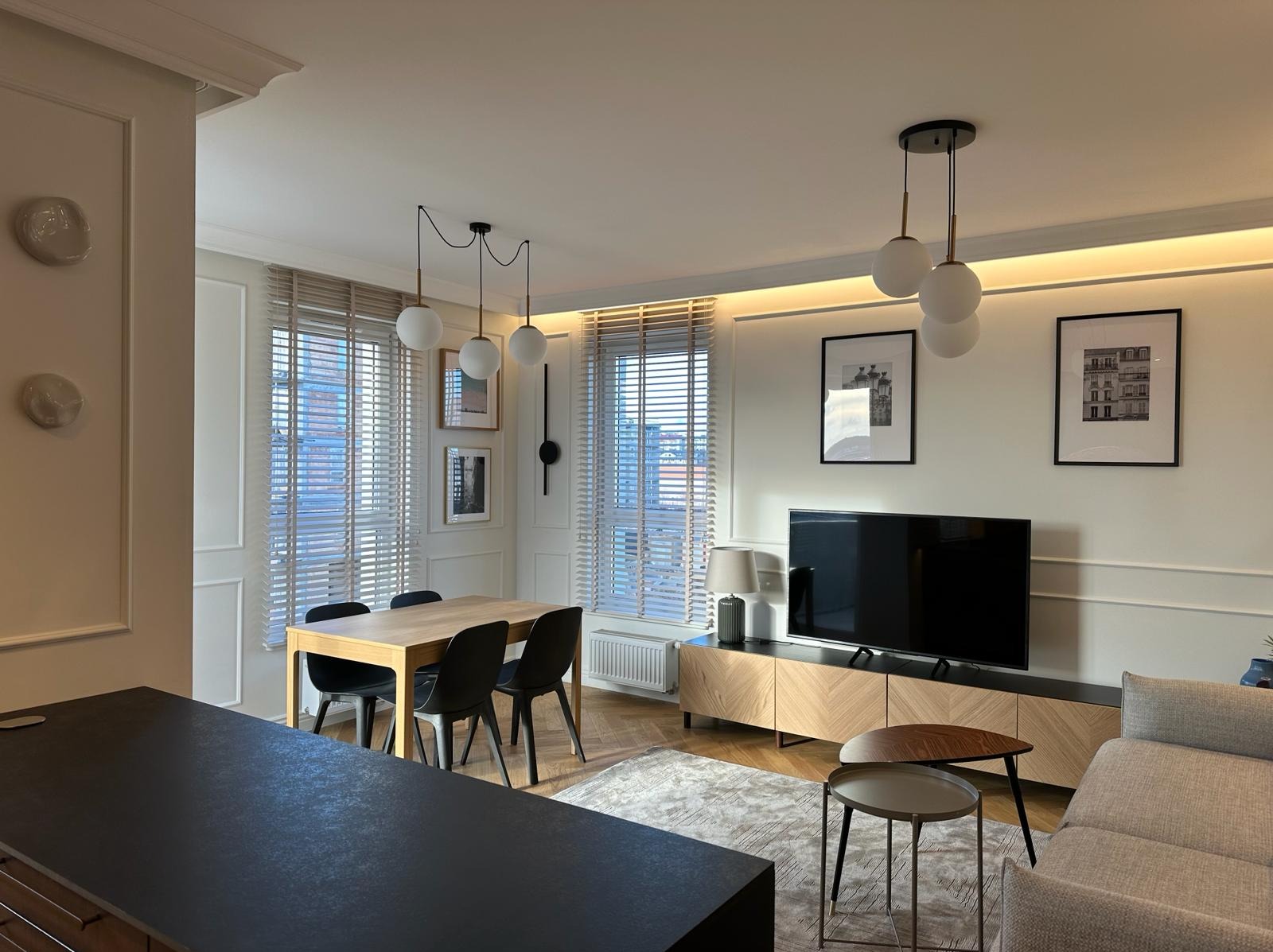 Jasne wnętrze salonu z jadalnią, drewniany stół z czarnymi krzesłami, telewizor na szafce RTV z forniru, białe ściany z dekoracyjnymi listwami, okna z jasnymi żaluzjami, lampy wiszące z trzema...