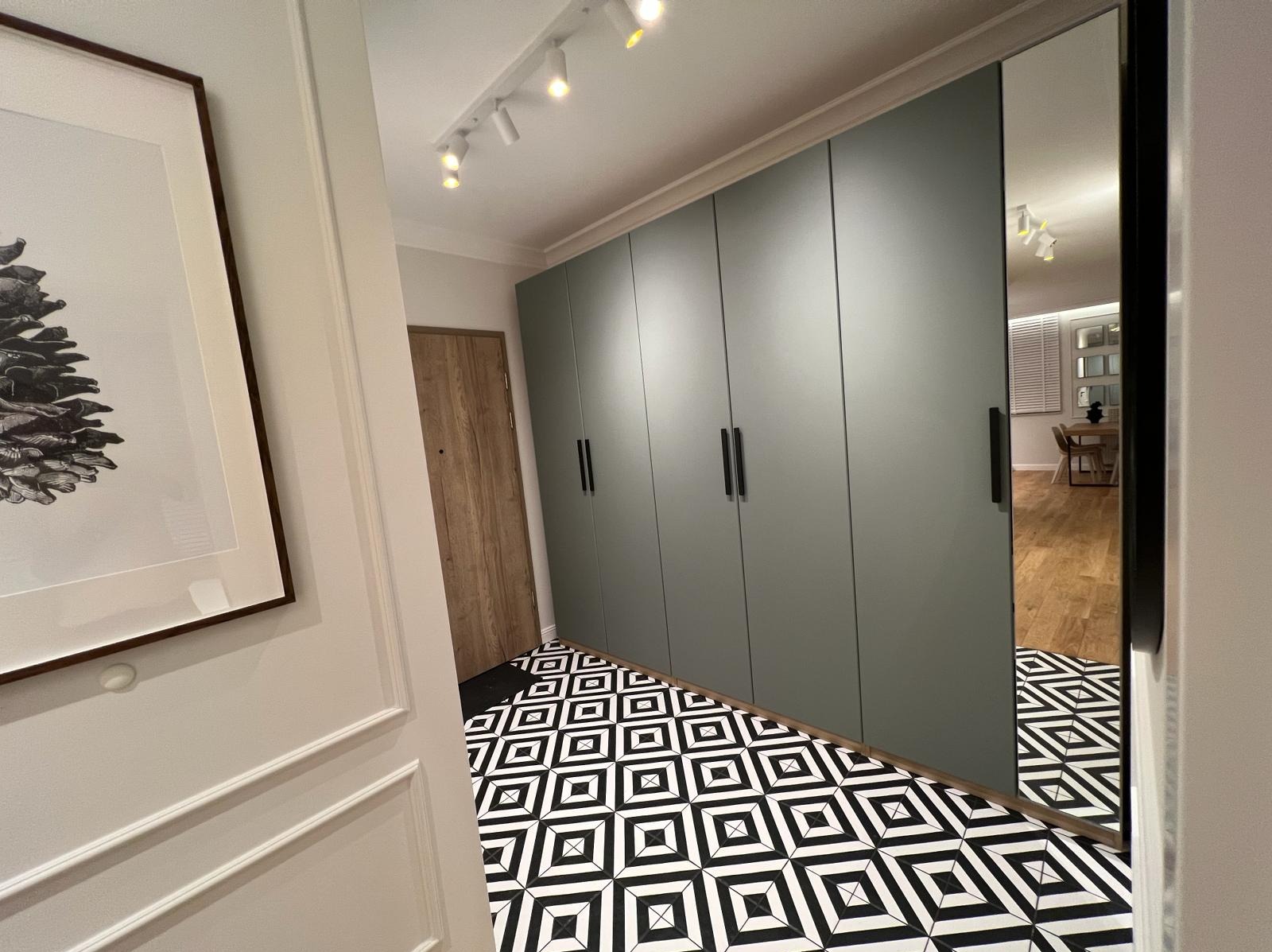 Wnętrze przedpokoju z dużą szafą w zabudowie w odcieniach szarości, wzorzystą czarno-białą podłogą, oświetleniem punktowym na suficie i lustrzanym frontem szafy odbijającym jadalnię.