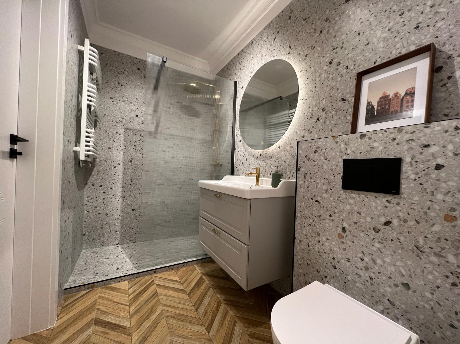 Nowoczesna łazienka w Gdyni z kabiną prysznicową, umywalką z szafką w kolorze gołębim, okrągłym lustrem z podświetleniem LED i płytkami imitującymi lastryko na ścianach oraz drewnopodobną podłogą...