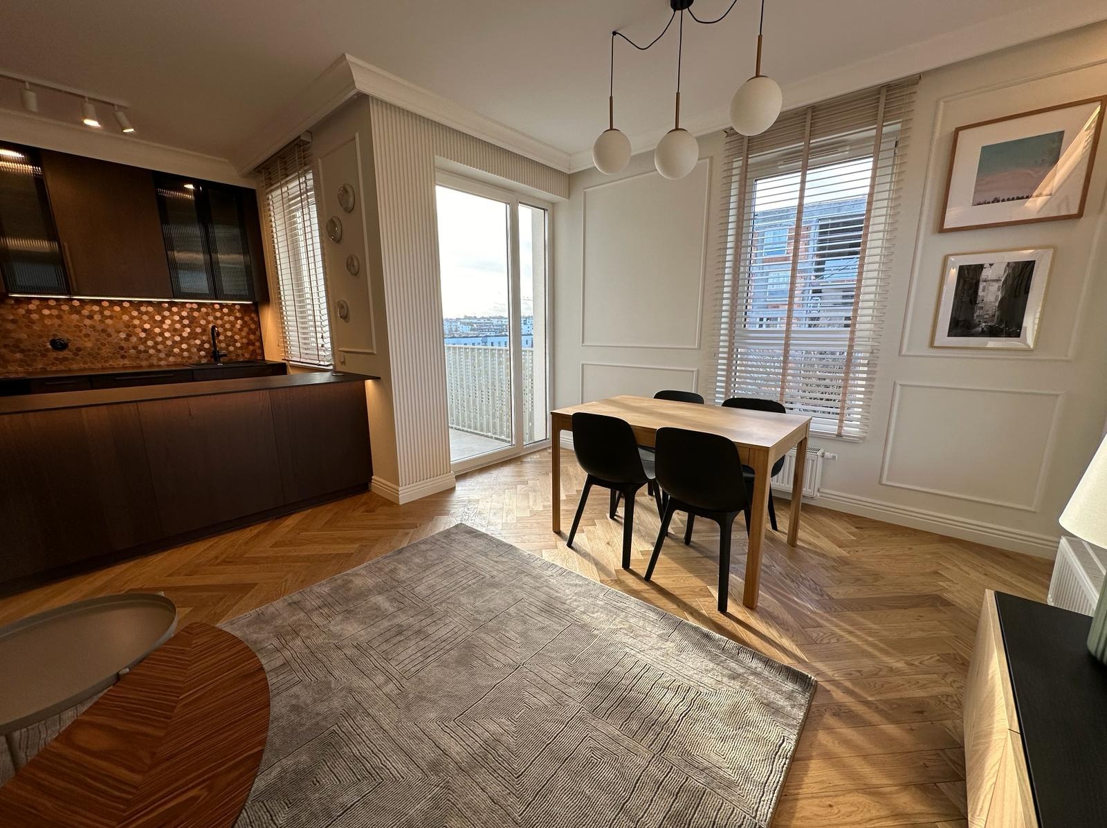 Jasne, przestronne wnętrze z parkietem w jodełkę, szarym dywanem w geometryczne wzory, drewnianym stołem z czarnymi krzesłami, nowoczesną kuchnią z mozaiką na ścianie i dużym oknem balkonowym...