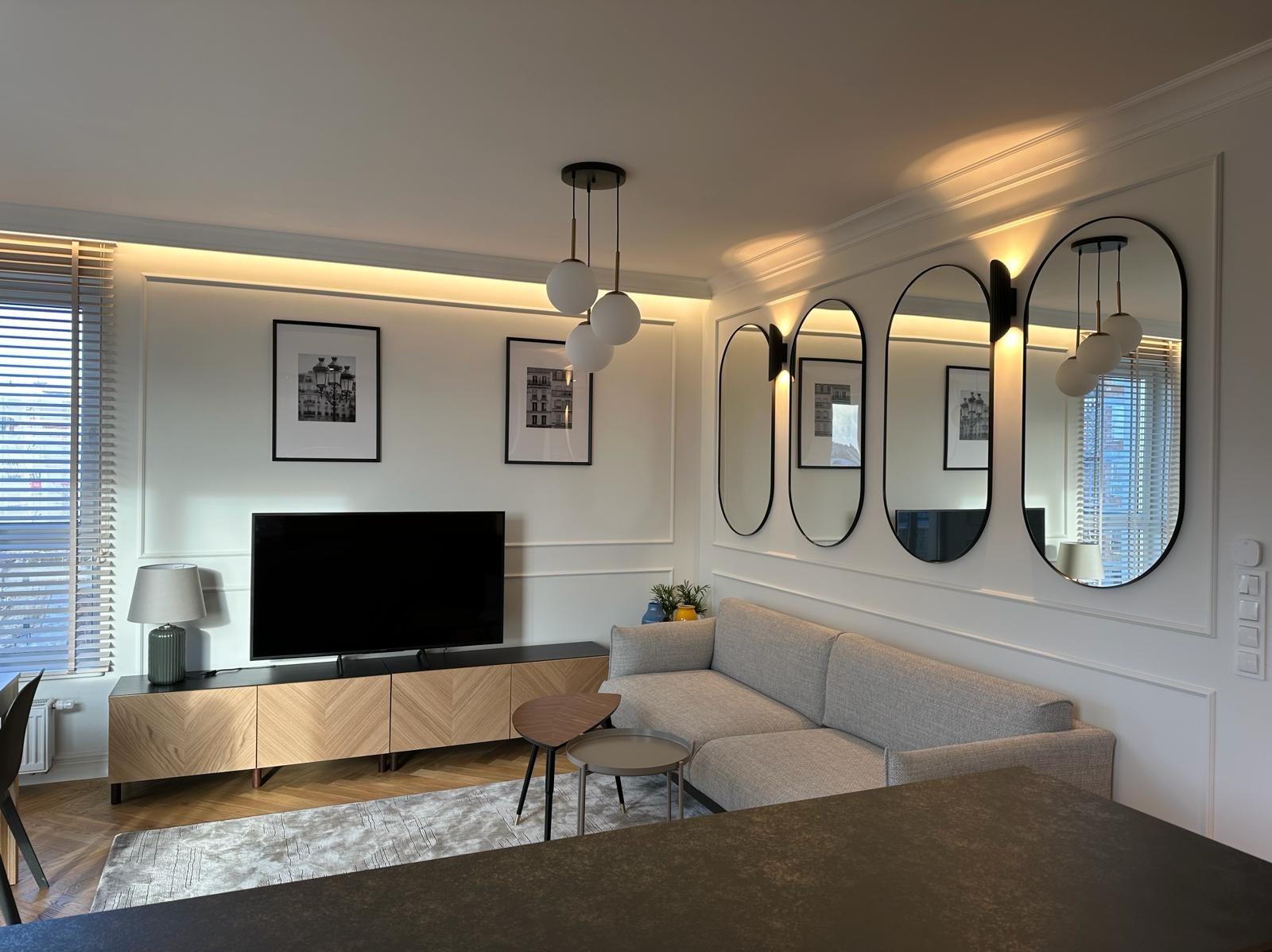Elegancki salon z szarą sofą narożną, trzema owalnymi lustrami z czarną ramą, podwieszanym oświetleniem LED, komodą z fornirem w jodełkę oraz dwoma stolikami kawowymi na dywanie w odcieniach szarości.