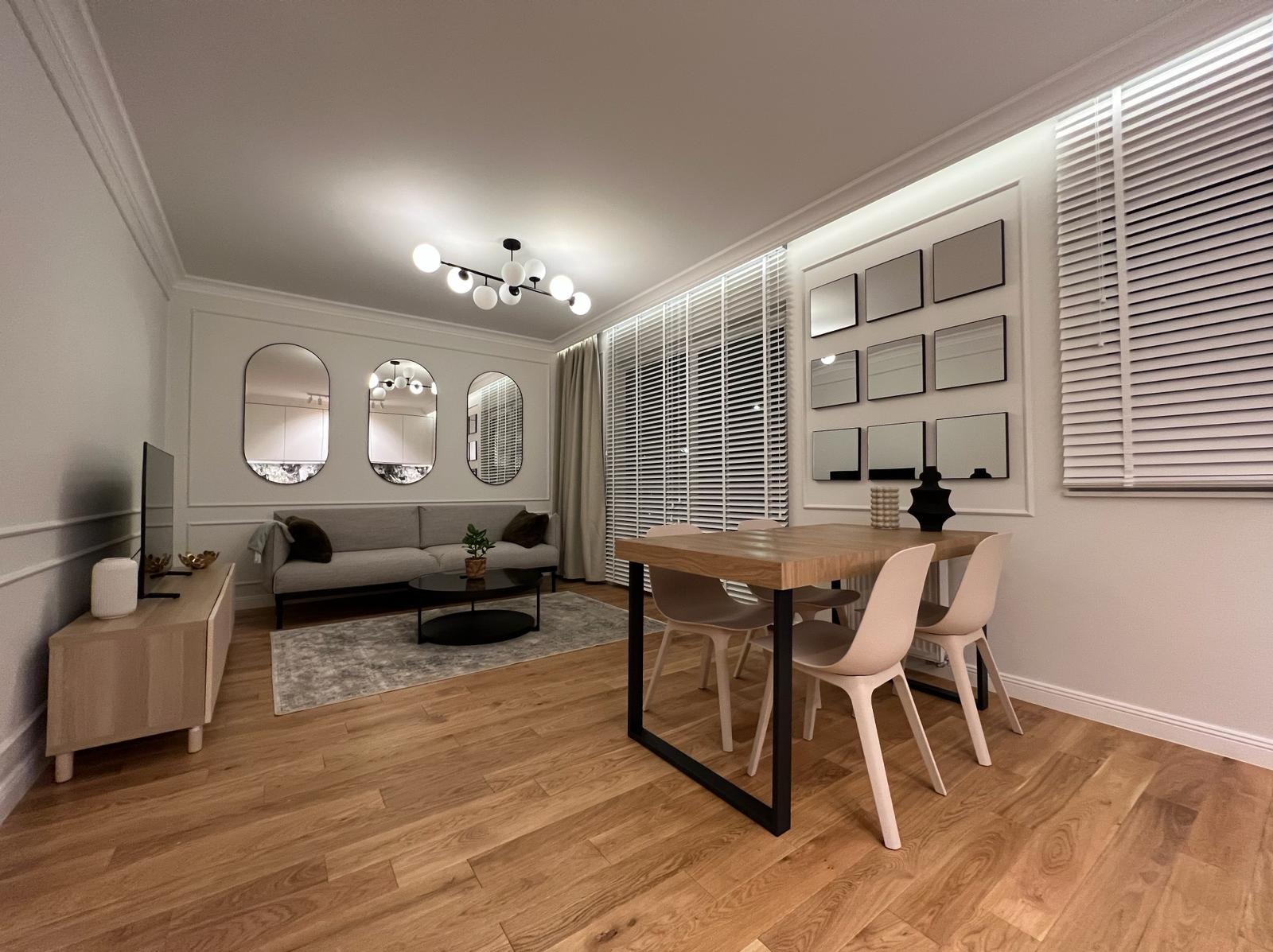 Jasne wnętrze salonu z drewnianą podłogą, szarą sofą, okrągłym stolikiem kawowym, stołem jadalnym z krzesłami i lustrami na ścianie.