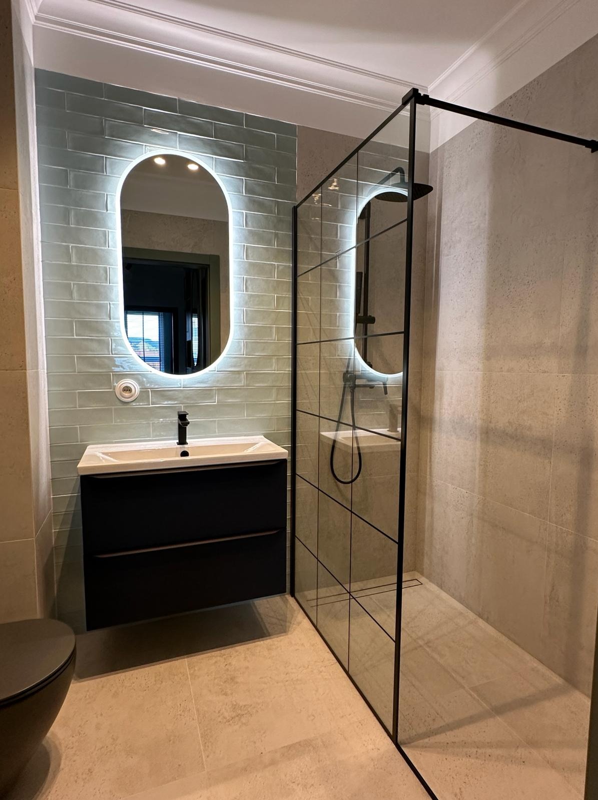 Nowoczesna łazienka z umywalką na szafce w kolorze czarnym, podświetlanym lustrem owalnym i kabiną prysznicową z czarnymi ramami na jasnych płytkach.