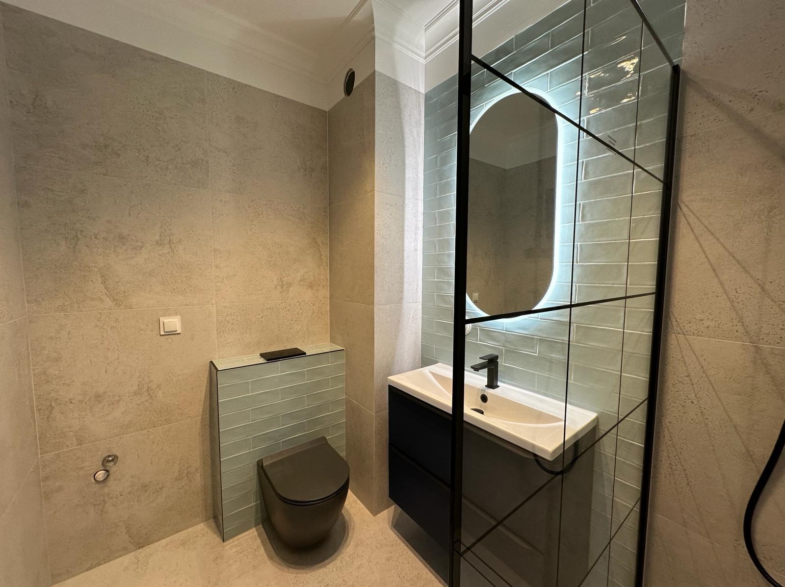 Nowoczesna łazienka z szarobeżowymi płytkami, podwieszaną toaletą i umywalką z czarną szafką, akcentem są niebieskie płytki za toaletą i podświetlane lustro w czarnej ramie.
