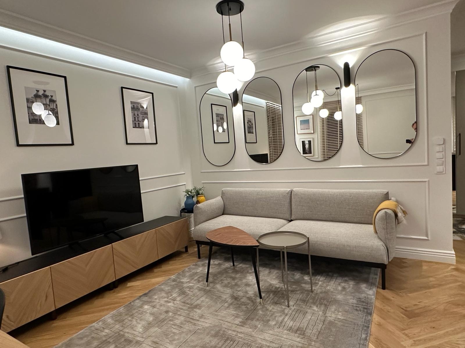 Nowoczesny salon z szarą sofą, stolikami kawowymi i owalnymi lustrami na ścianie z dekoracyjną sztukaterią, oświetlony lampą wiszącą z kulistymi kloszami.