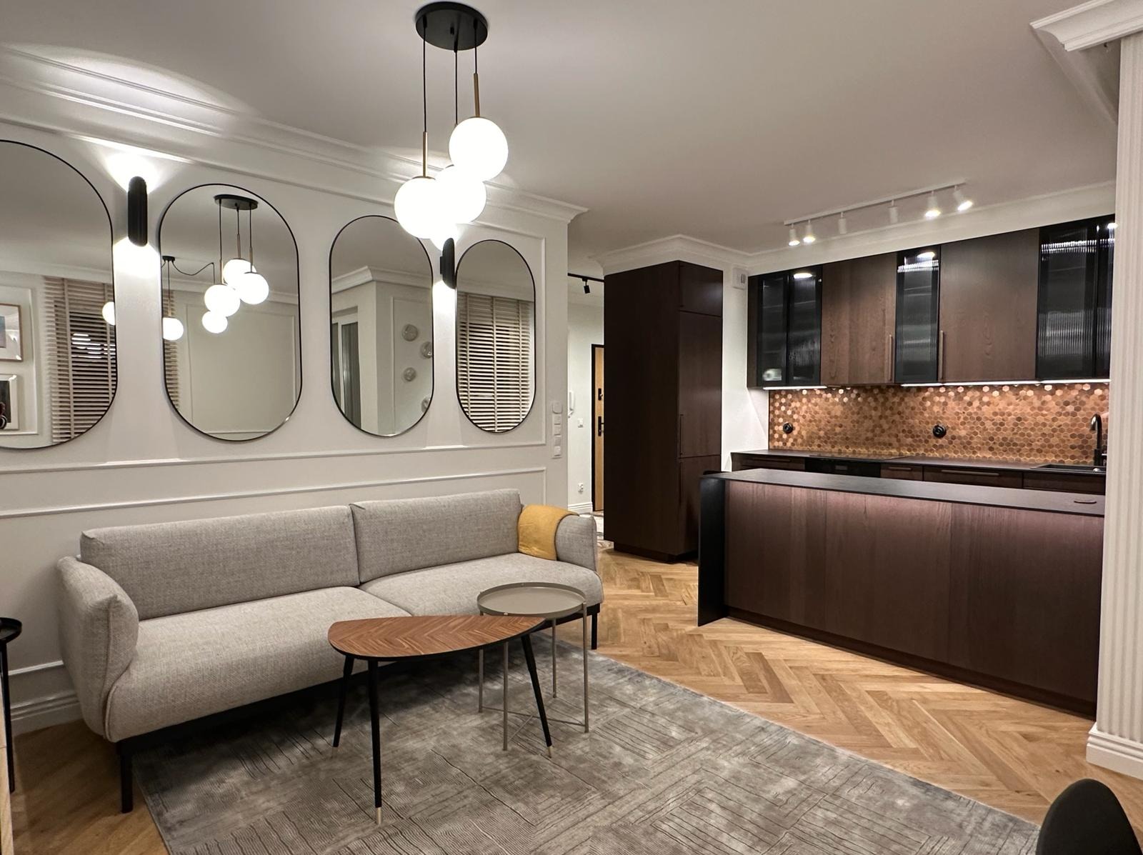 Salon z szarą sofą, stolikami kawowymi, trzema owalnymi lustrami na ścianie z dekoracyjną sztukaterią i widoczną w tle zabudową kuchenną z drewnianymi frontami i mozaiką w odcieniach miedzi.