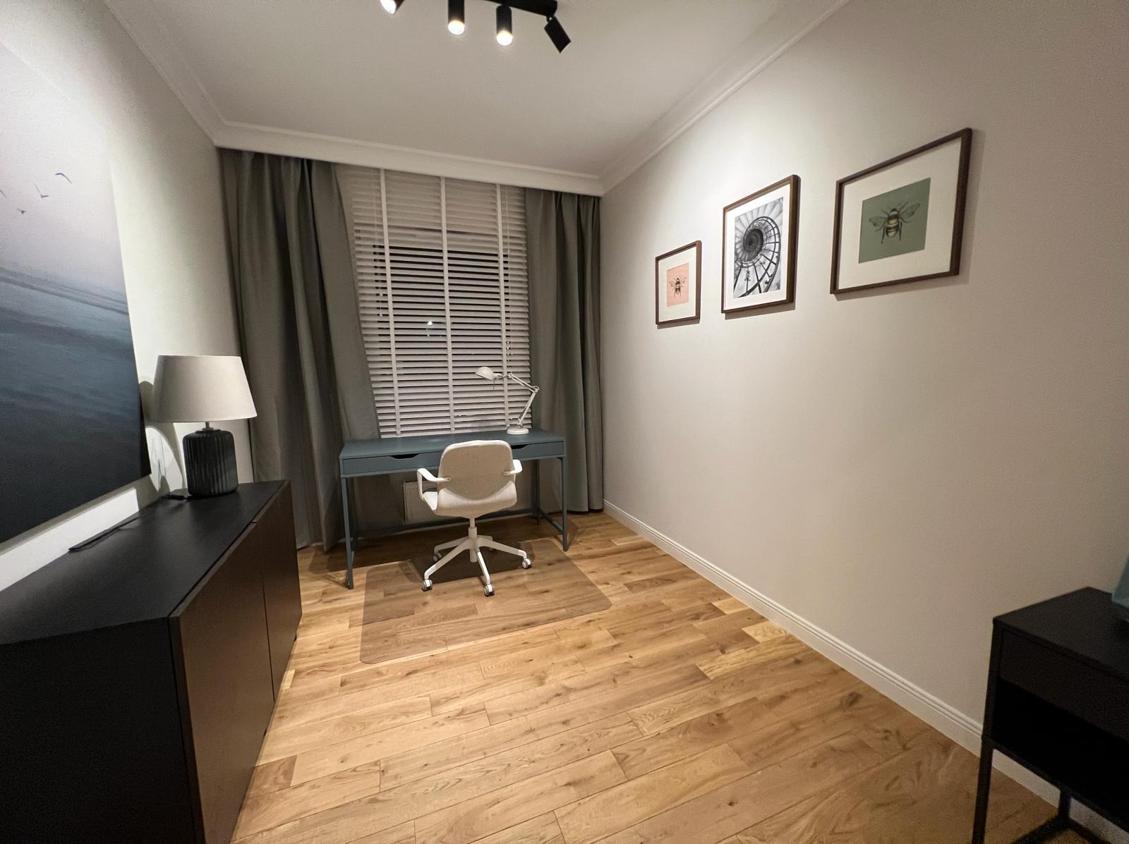 Pokój z drewnianą podłogą, biurkiem z krzesłem na kółkach, lampką, zasłonami i trzema obrazkami na ścianie. Na podłodze przezroczysta mata ochronna.