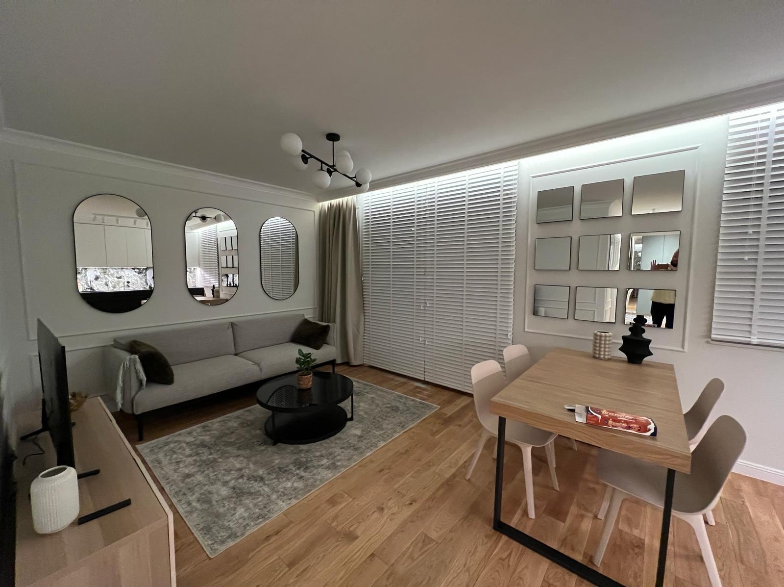 Jasne wnętrze salonu z drewnianą podłogą, szarą sofą, okrągłym stolikiem kawowym, zasłonami, żaluzjami, trzema owalnymi lustrami na ścianie i stołem z krzesłami.
