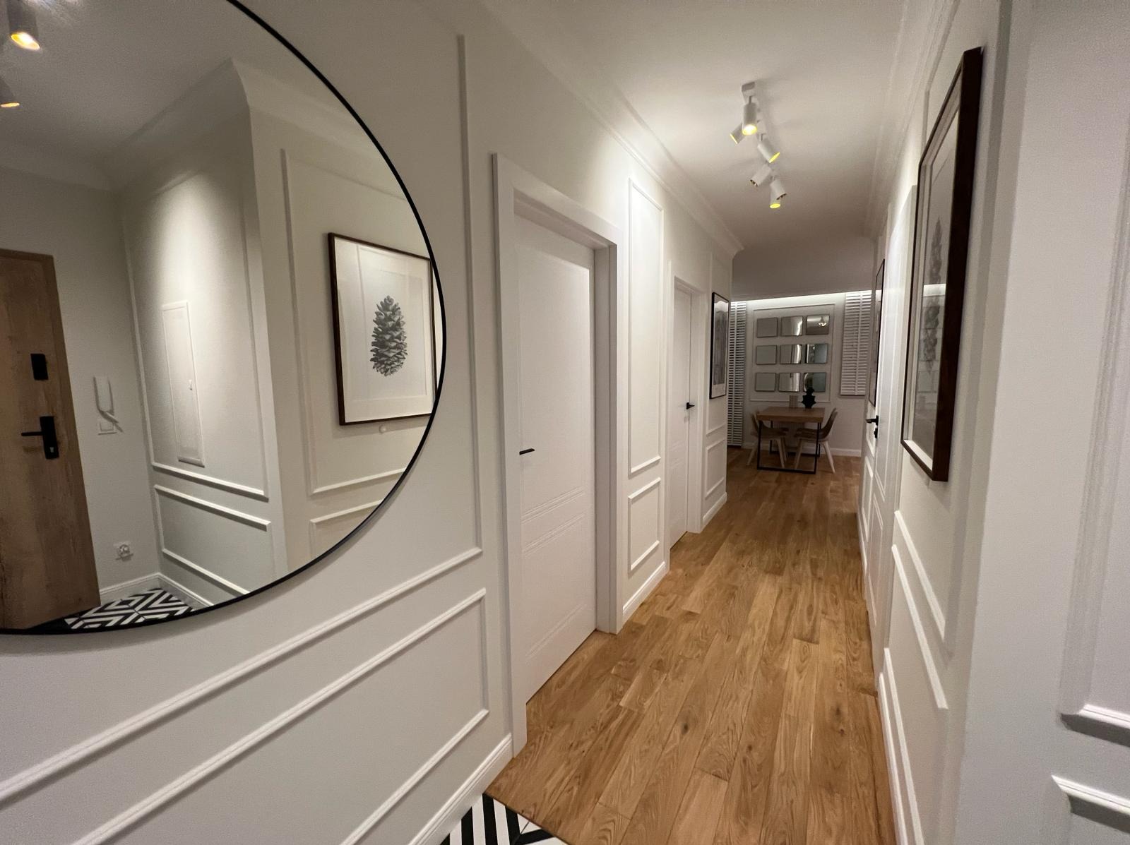 Nowoczesny korytarz z drewnianą podłogą, białymi ścianami z dekoracyjnymi listwami, okrągłym lustrem i widokiem na jadalnię w głębi.