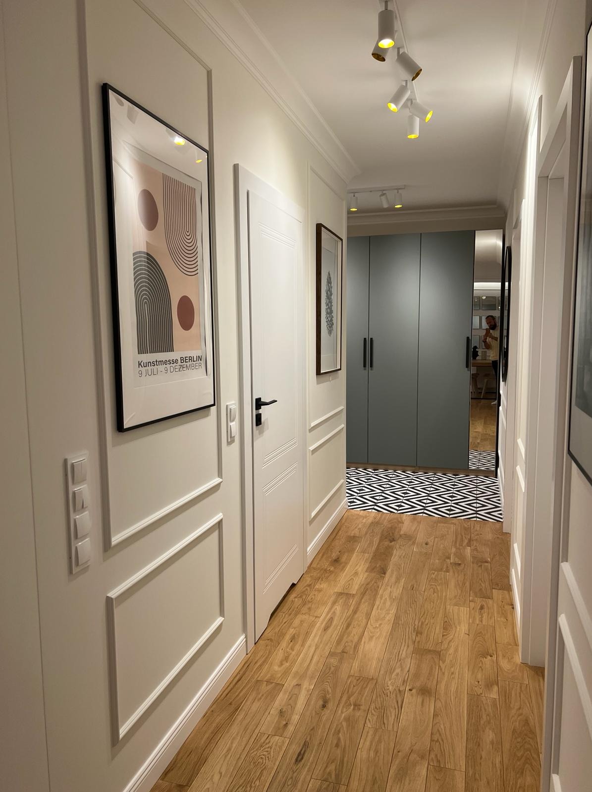 Korytarz z drewnianą podłogą, białymi ścianami z dekoracyjnymi listwami, białymi drzwiami z czarnymi klamkami, nowoczesnymi obrazami na ścianach i szafą w zabudowie w tle.