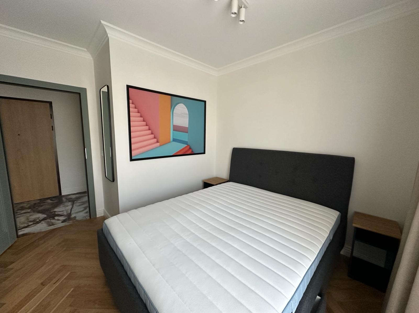 Nowoczesna sypialnia z parkietem w jodełkę, łóżkiem z szarą ramą i zagłówkiem, białą pościelą oraz abstrakcyjnym obrazem w czarnej ramie wiszącym nad łóżkiem.