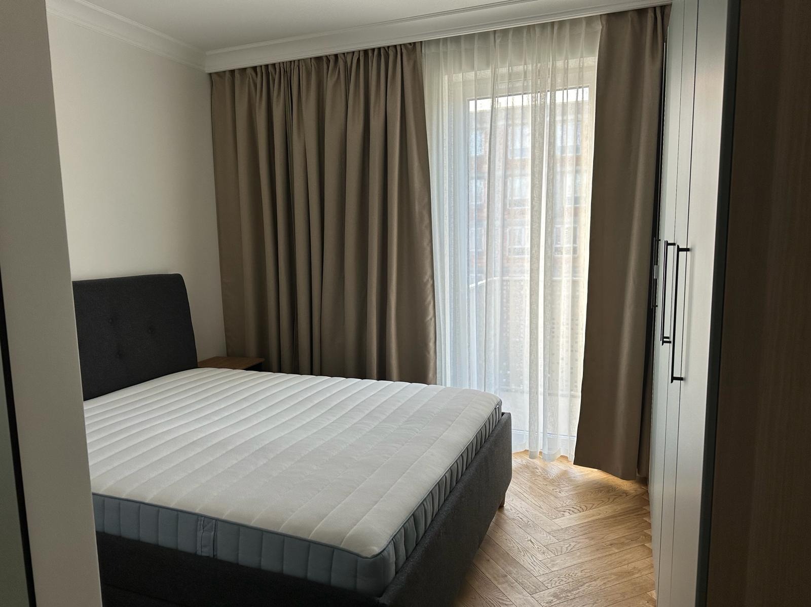 Sypialnia z łóżkiem kontynentalnym, parkietem w jodełkę i zasłonami w odcieniach brązu, widok na okno z firanką i fragment szafy z czarnymi uchwytami.