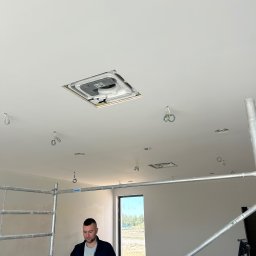 Concept4Energy Arkadiusz Godek - Instalator trzyma panel klimatyzacyjny na tle rusztowania w pomieszczeniu z otworami w suficie przygotowanymi pod montaż klimatyzacji kasetonowej.