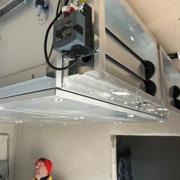 Concept4Energy Arkadiusz Godek - Szara, metalowa centrala wentylacyjna z czarnymi, obrotowymi elementami zamontowana pod sufitem, widoczny pracownik w czerwonej czapce spoglądający w górę.