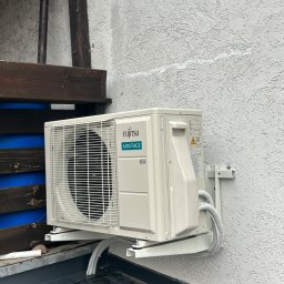 Concept4Energy Arkadiusz Godek - Zewnętrzna jednostka klimatyzacji Fujitsu Airstage zamontowana na wspornikach przy teksturowanej, jasnej ścianie budynku; widoczne przewody w peszlach i fragment dachu pokrytego blachą na rąbek...