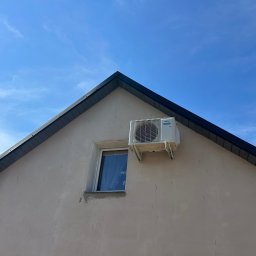 Concept4Energy - Biała jednostka zewnętrzna klimatyzatora marki Fujitsu zamontowana na elewacji domu obok okna, na tle błękitnego nieba z delikatnymi chmurami.