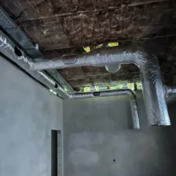 Szczegółowe ujęcie instalacji wentylacyjnej z aluminiową izolacją w surowym wnętrzu budynku w trakcie budowy, widoczne kable i izolacja stropu.