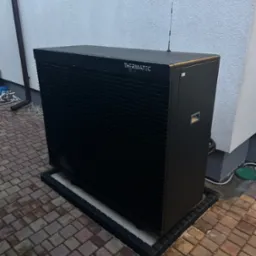 Czarna pompa ciepła marki Thermatec Line zainstalowana na zewnątrz budynku, widoczna antena na górze urządzenia, kostka brukowa wokół.