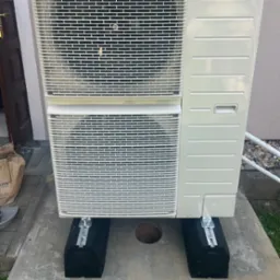 Zewnętrzna jednostka klimatyzacji umieszczona na czarnych podkładkach z aluminiowymi szynami, widoczny otwór w betonowej płycie, rury instalacyjne w tle.