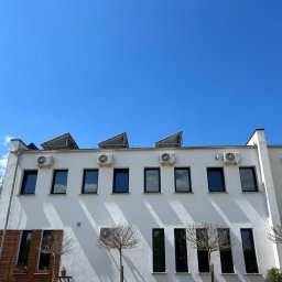 Concept4Energy Arkadiusz Godek - Biały budynek z rzędem okien i klimatyzatorów zamontowanych na elewacji pod dachem, z panelami słonecznymi na dachu, na tle błękitnego nieba.