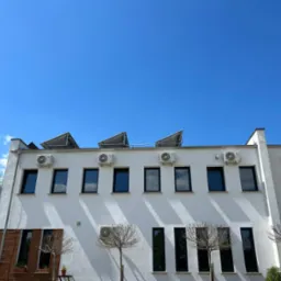 Biały budynek z rzędem okien i klimatyzatorów zamontowanych na elewacji pod dachem, z panelami słonecznymi na dachu, na tle błękitnego nieba.
