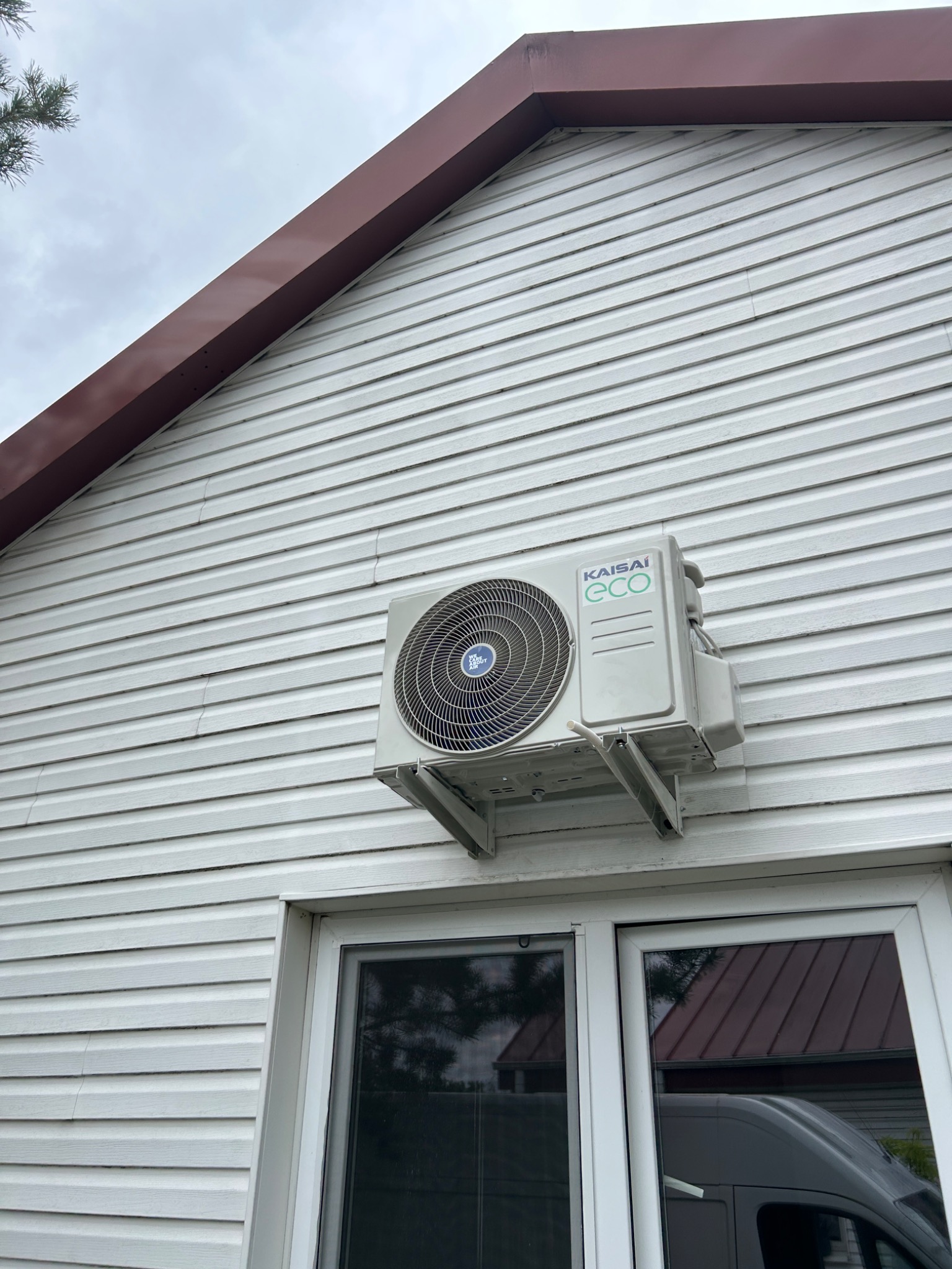 Zewnętrzna jednostka klimatyzacji Kaisai Eco zamontowana na białej elewacji domu z sidingu, widoczna nad oknem z odbiciem samochodu dostawczego.