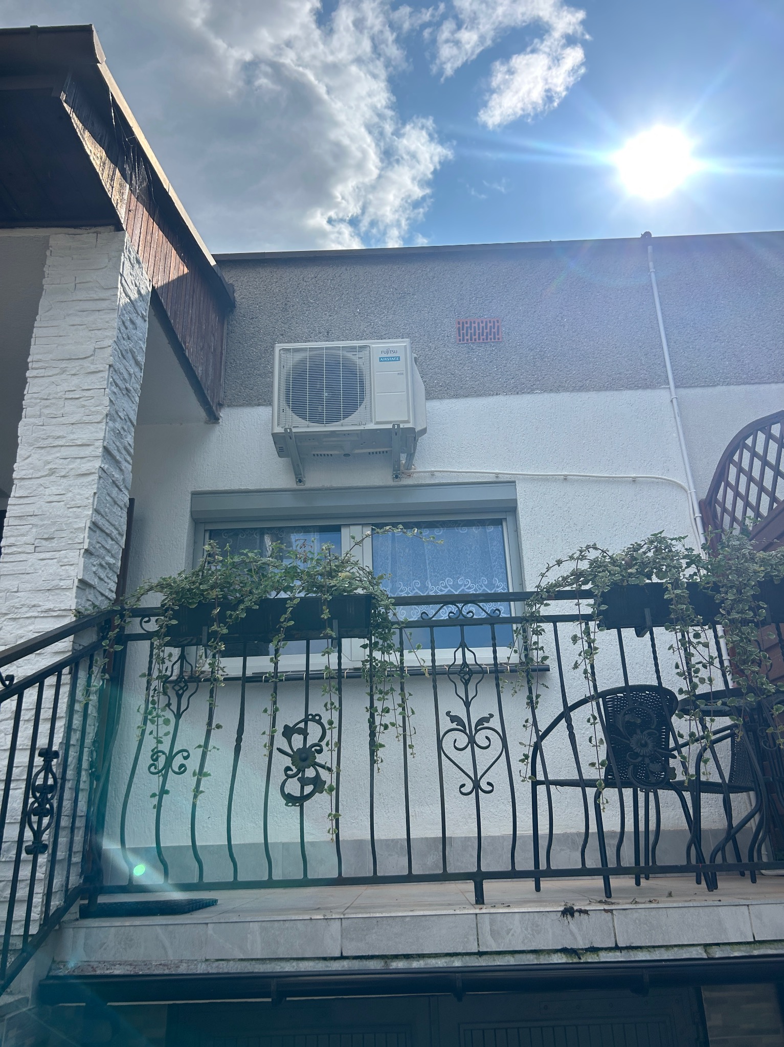 Biała jednostka zewnętrzna klimatyzatora zamontowana na ścianie budynku z balkonem, widoczna z dołu, z promieniami słońca w kadrze.